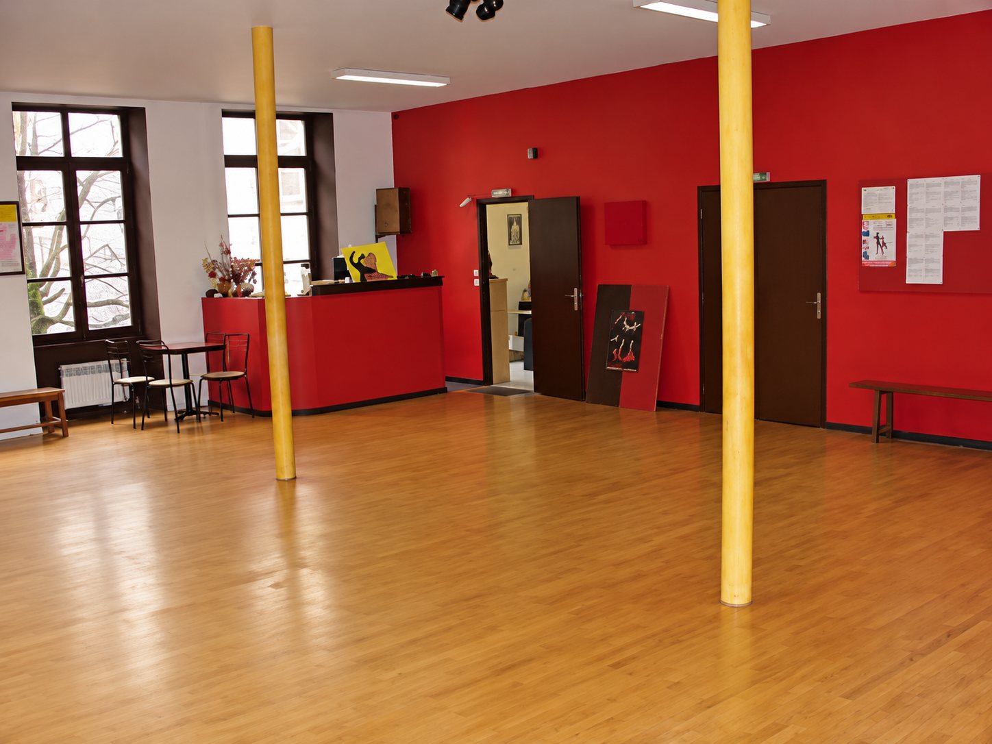 Une grande salle vide avec un parquet en bois clair, des murs rouges, deux poteaux en bois et un comptoir rouge au fond.
