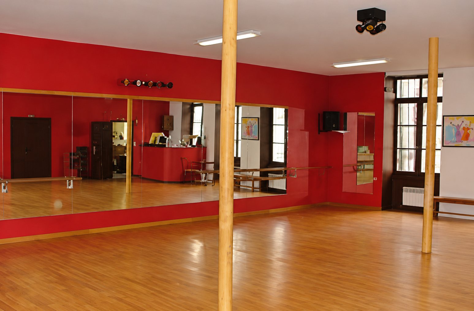 Une salle de danse lumineuse aux murs rouges, dotée d'un grand miroir, d'un parquet en bois clair et de deux poteaux verticaux.