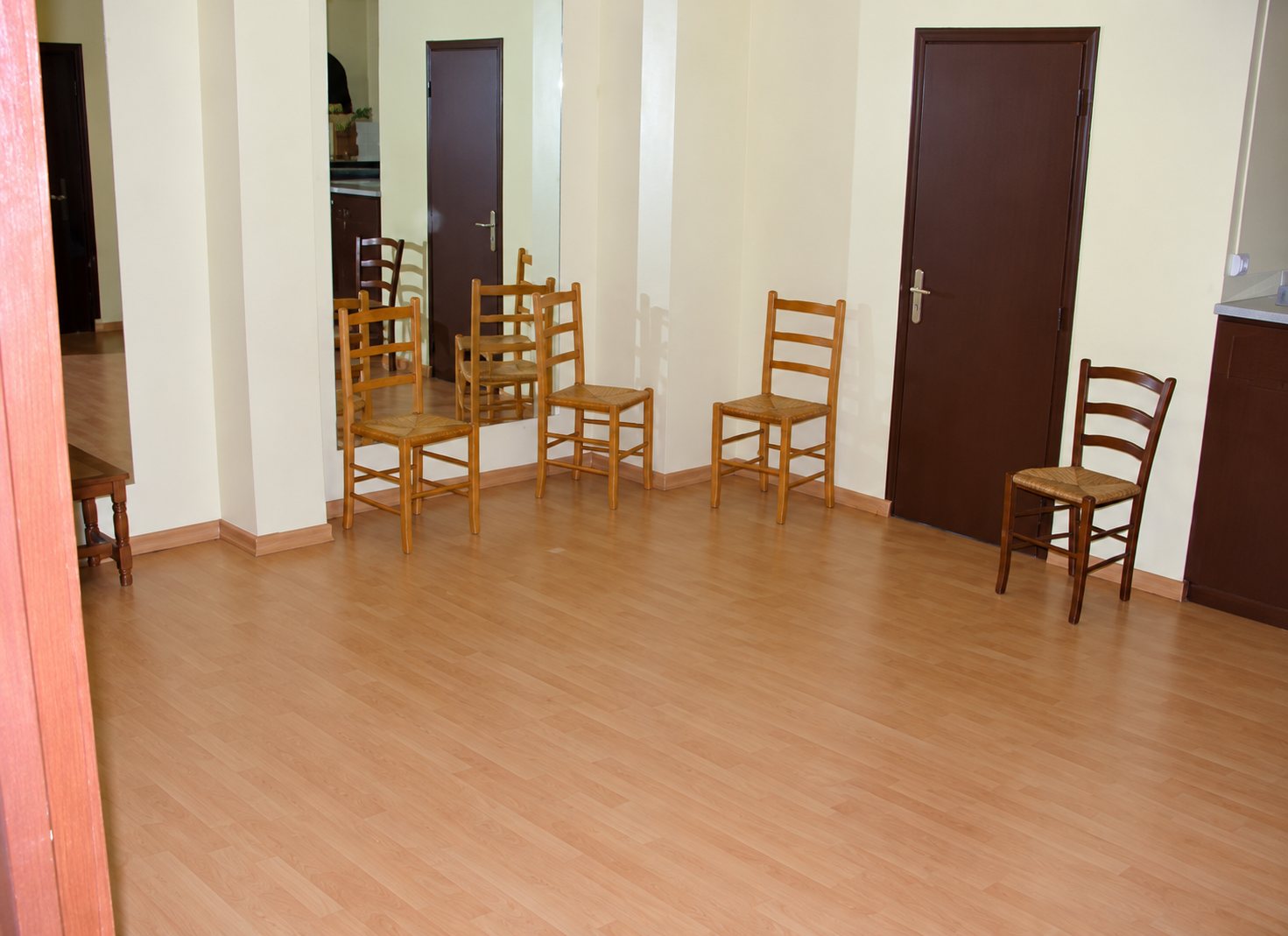 Une pièce vide au sol en parquet clair, avec plusieurs chaises en bois à assise paillée disposées le long des murs jaunes près de portes brunes.